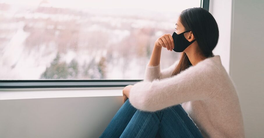 woman with face mask looking outside the window on winter