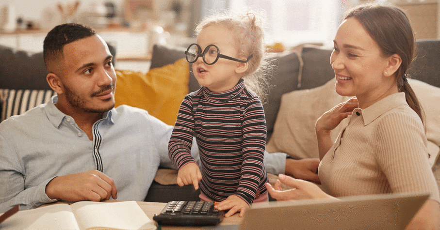 Portrait of happy young family counting home finances with cute little girl wearing glasses in sunlit apartment