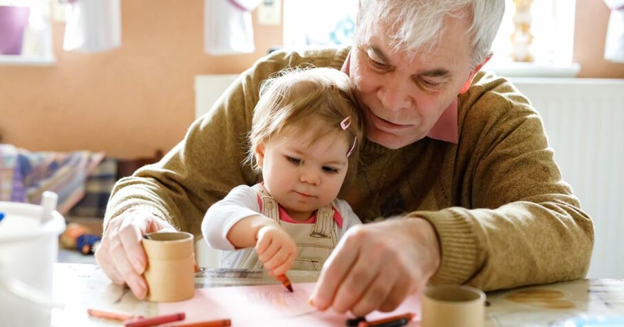 Cute little baby toddler girl and handsome senior grandfather painting with colorful pencils at home. Grandchild and man having fun together