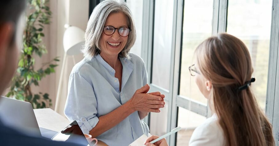 Happy mature old female mentor coach laughing training young interns at group office meeting professional workshop. Cheerful middle aged teacher professor talking to students at university