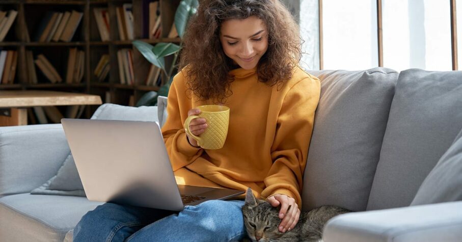 Happy relaxed hispanic teen girl student or freelancer relaxing sitting on couch holding laptop on lap working studying from home drinking tea, playing with cute pet cat in cozy sunny room on sofa.