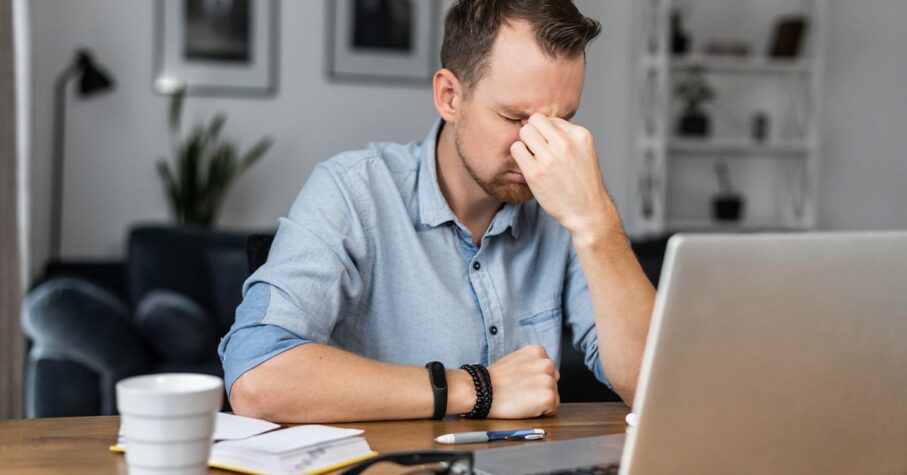 Overworked young entrepreneur in tired from work with a laptop, holds on to the bridge of the nose his eyes closed. Stressed guy works remotely at home feels eyes strain