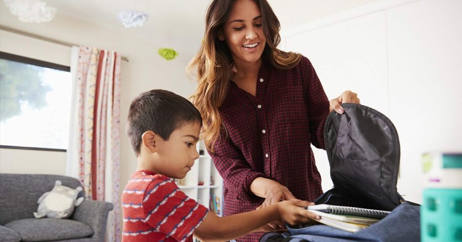 Mother In Bedroom Helping Son To Pack Bag Ready For School