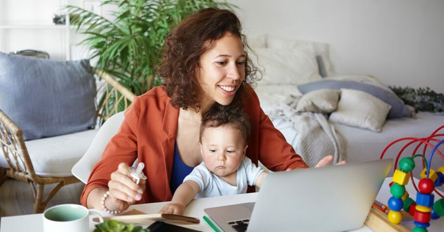 woman working at desk at home using laptop, holding baby on her lap